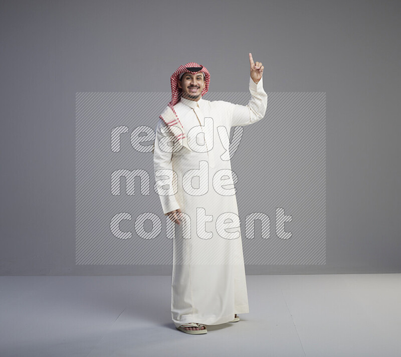 A Saudi man standing wearing thob and red shomag interacting with the camera on gray background