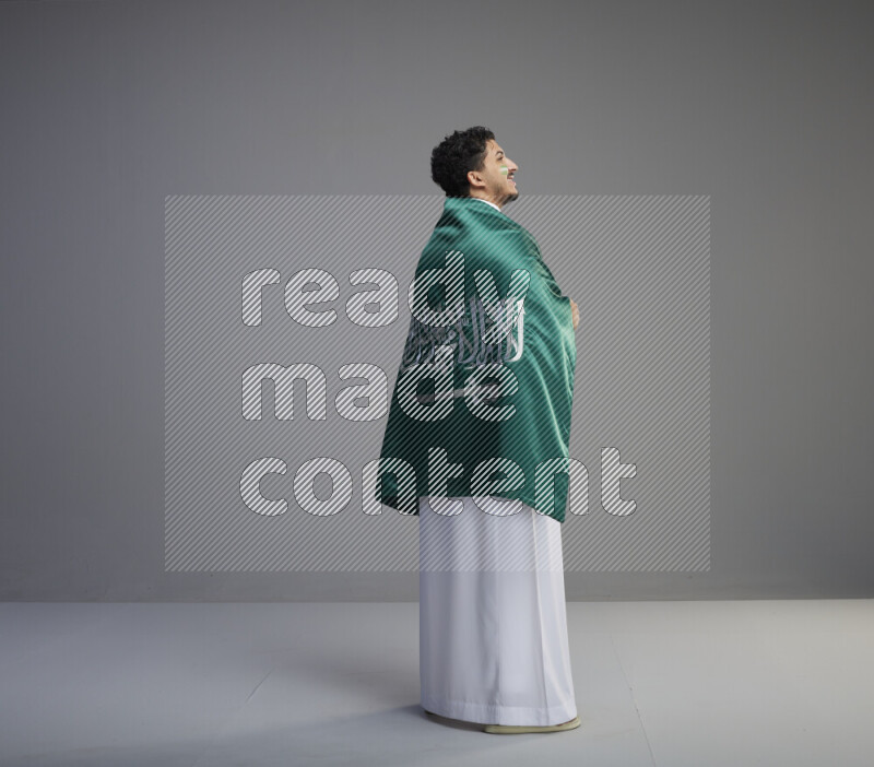 A Saudi man standing wearing thob with face painting wrapping big Saudi flag on gray background