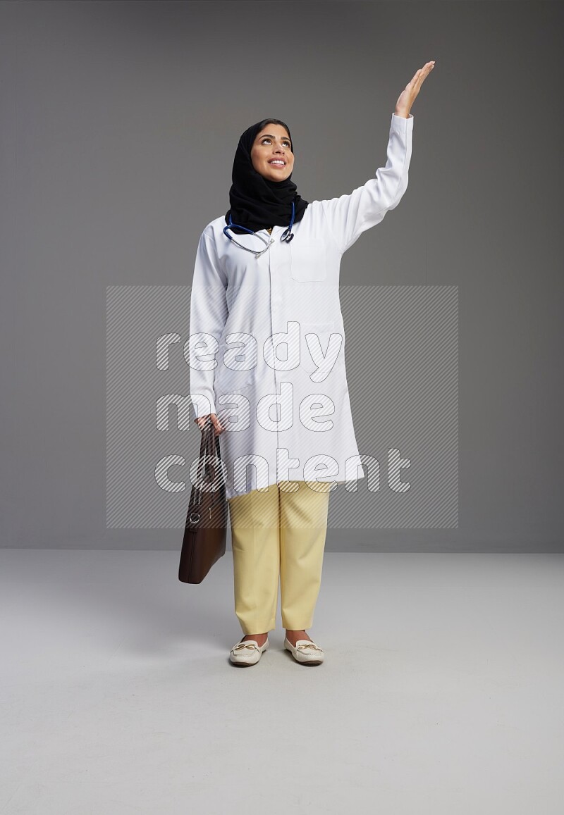 Saudi woman wearing lab coat with stethoscope standing holding bag on Gray background