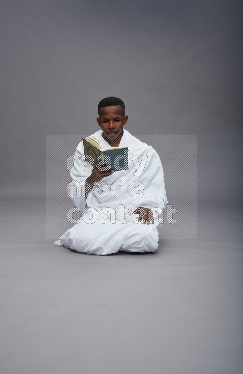 A man wearing Ehram sitting on floor reading quran on gray background