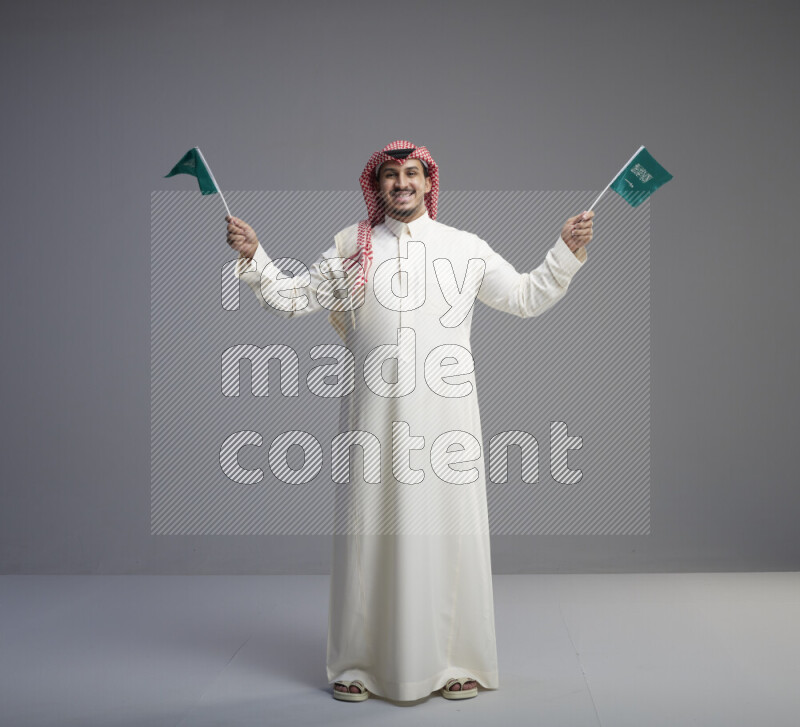 A Saudi man standing wearing thob and red shomag raising small Saudi flag on gray background