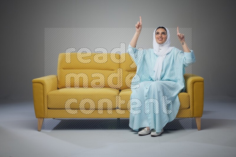 A Saudi woman wearing a light blue Abaya and white head scarf sitting on a yellow sofa and thinking and pointing up eye level on a grey background