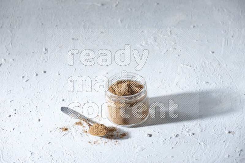 A glass jar and a metal spoon full of allspice powder on a textured white flooring
