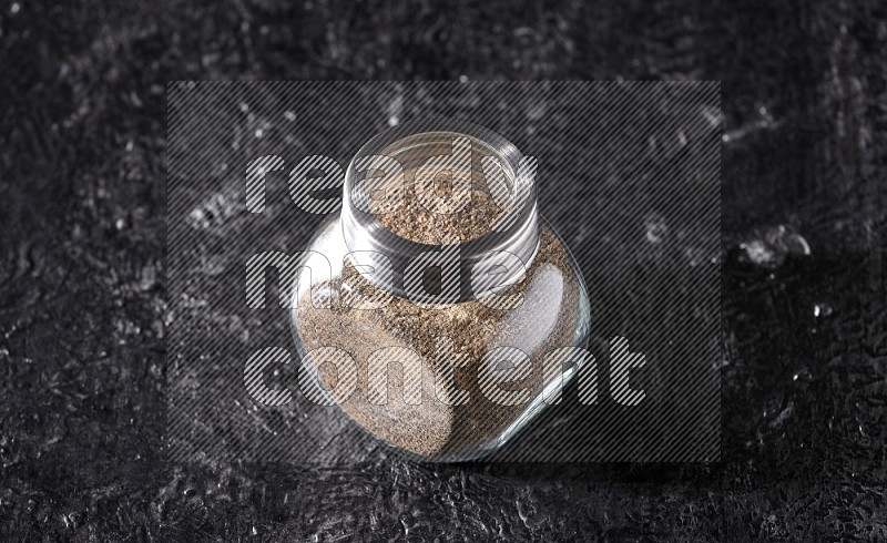A glass spice jar full of black pepper powder on textured black flooring
