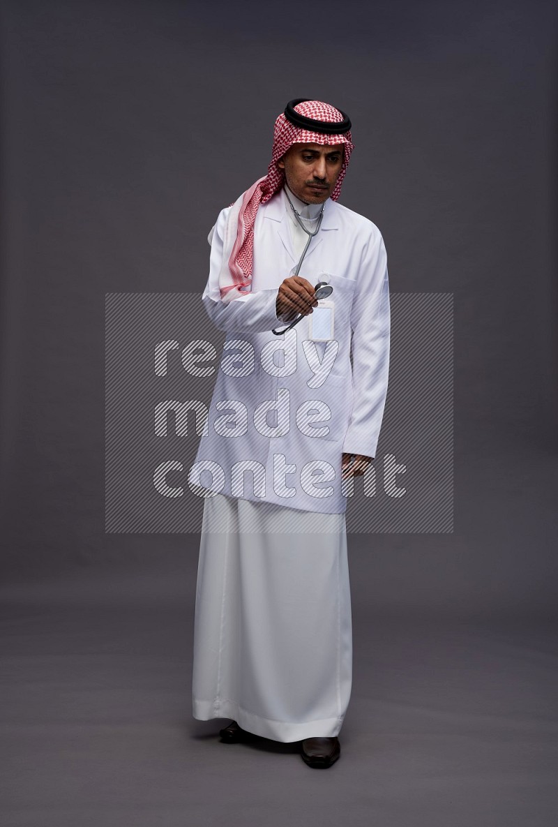 Saudi man wearing thob with lab coat and shomag with pocket employee badge with stethoscope standing interacting with the camera on gray background