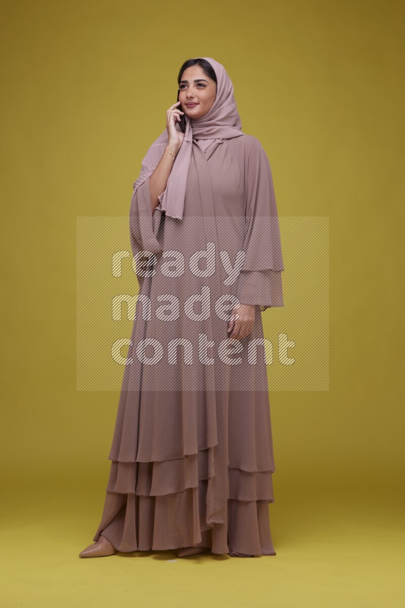 A woman having a call on a Yellow Background wearing Brown Abaya with Hijab