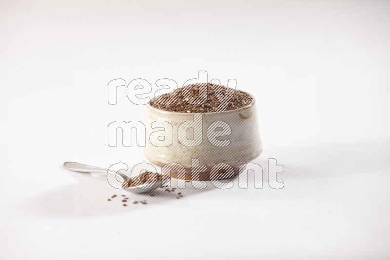 A pottery beige bowl and a metal spoon full of flax seeds on a white flooring