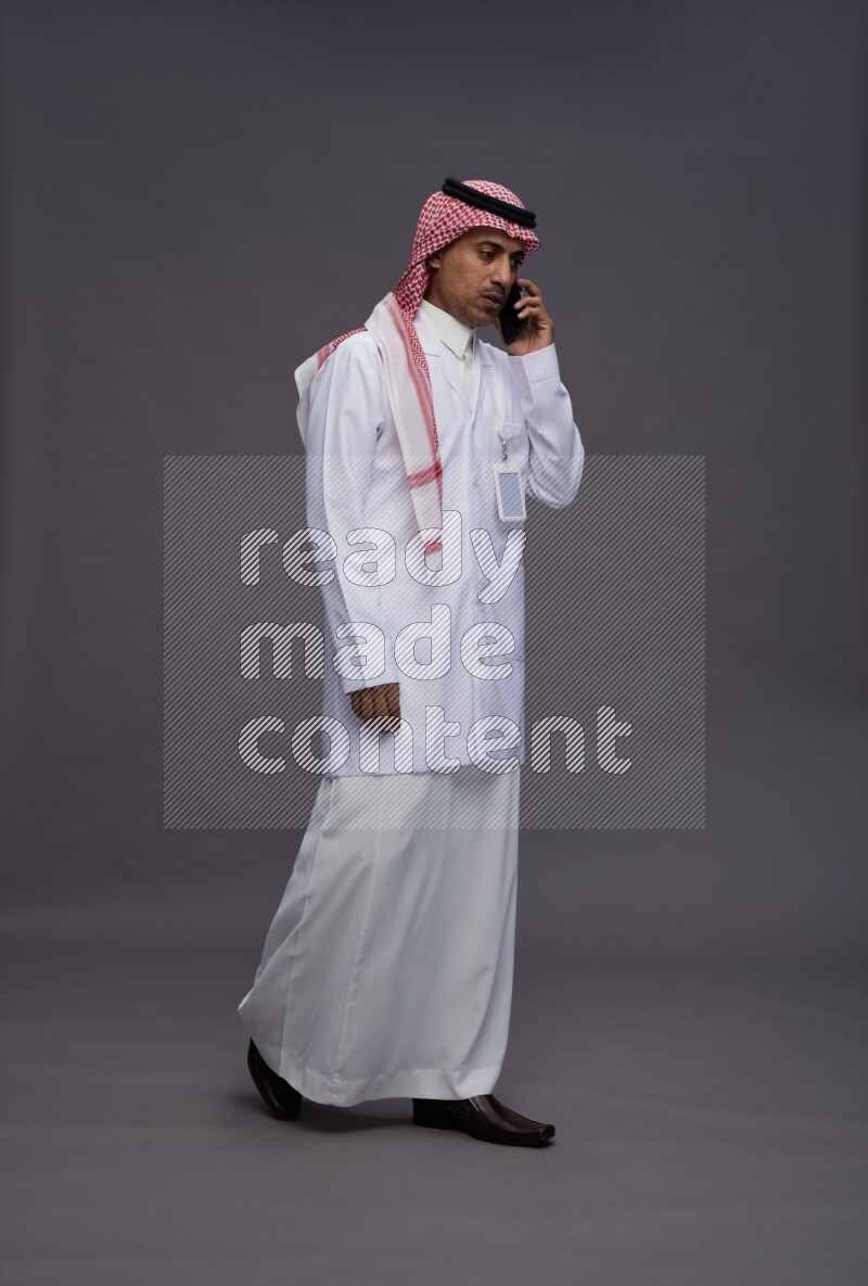 Saudi man wearing thob with lab coat and shomag with pocket employee badge standing talking on phone on gray background