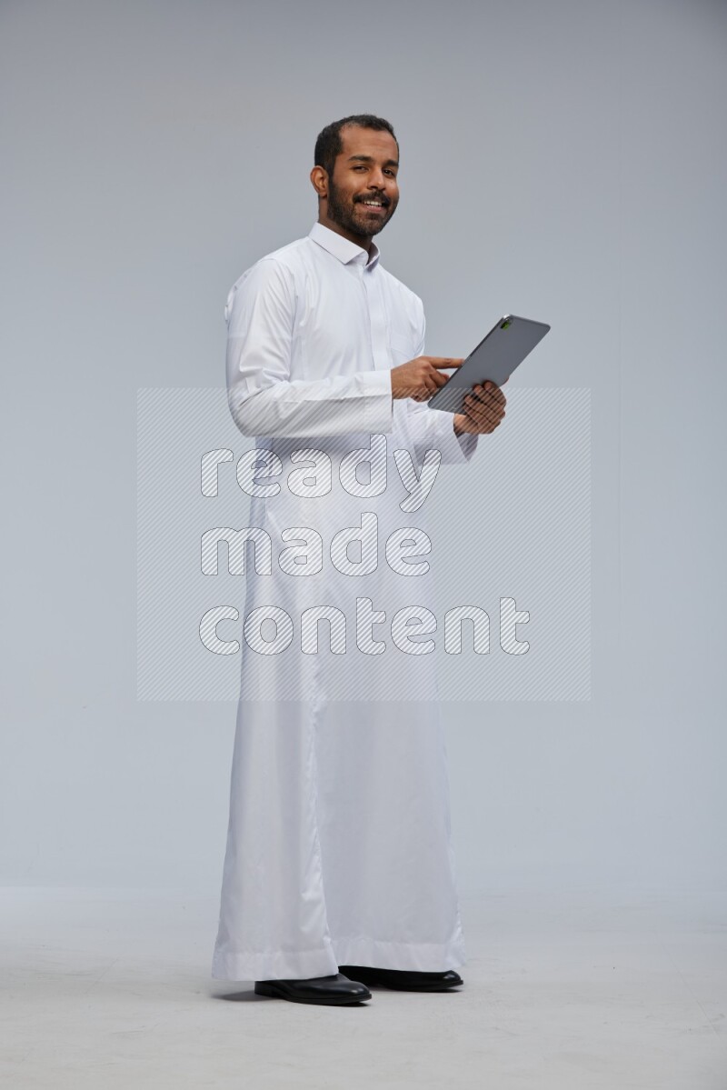 Saudi man wearing Thob standing working on tablet on Gray background