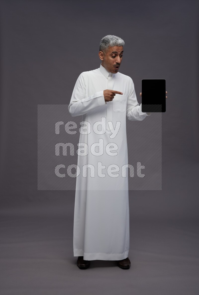 Saudi man wearing thob standing showing tablet to camera on gray background