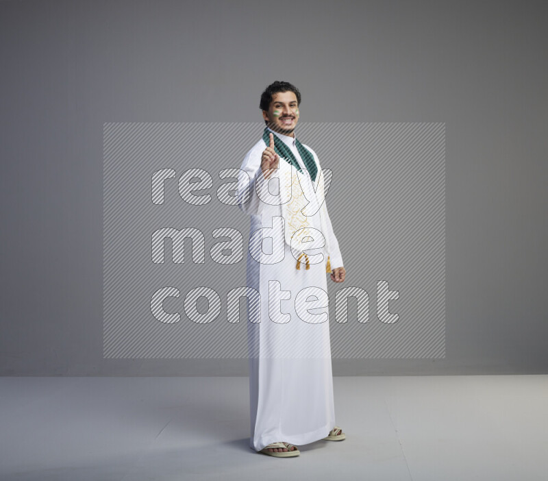 A Saudi man standing wearing thob and Saudi flag scarf with face painting on gray background