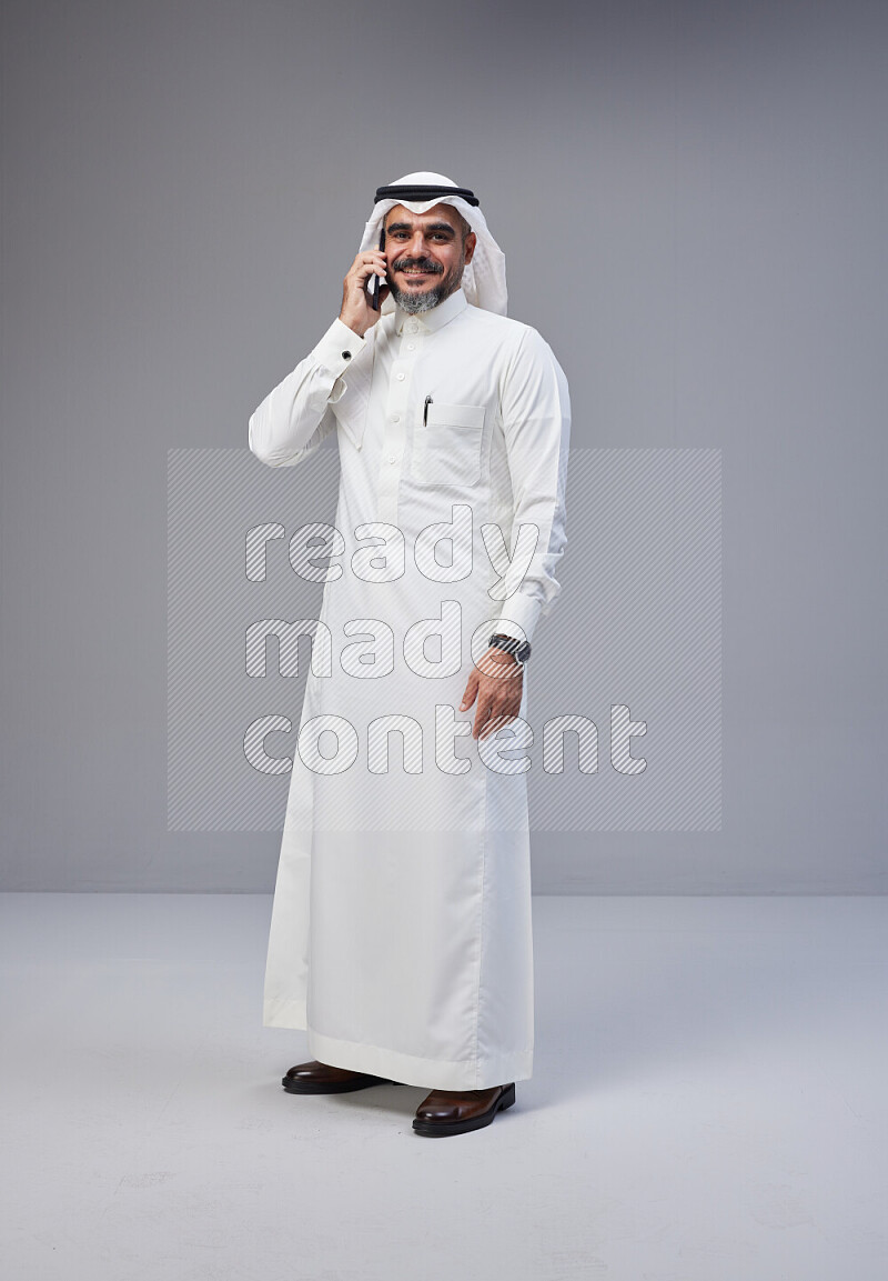 Saudi man Wearing Thob and white Shomag standing talking on phone on Gray background