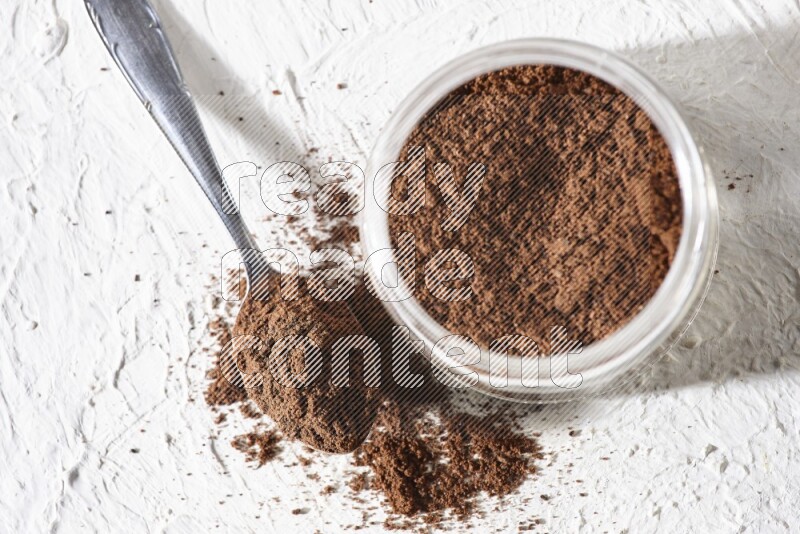 A glass jar full of cloves powder with a metal spoon on a textured white flooring