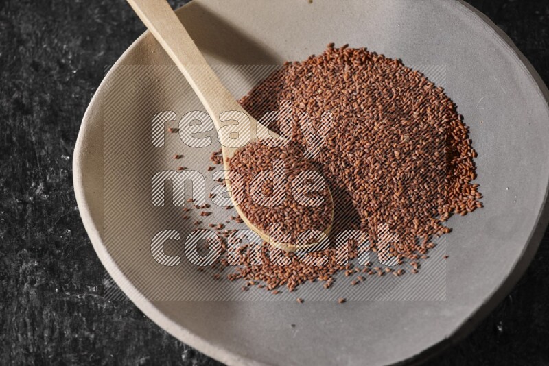 A multicolored pottery plate full of garden cress seeds and wooden spoon full of seeds on a textured black flooring