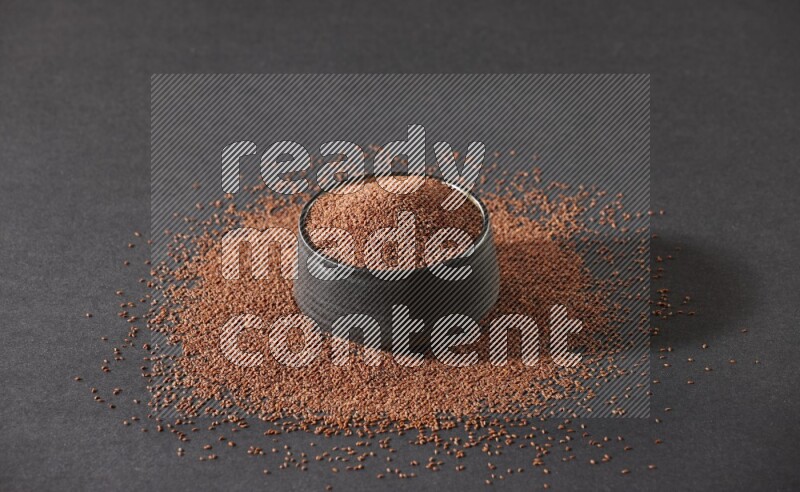 A black pottery bowl full of garden cress seeds surrounded by seeds on a black flooring