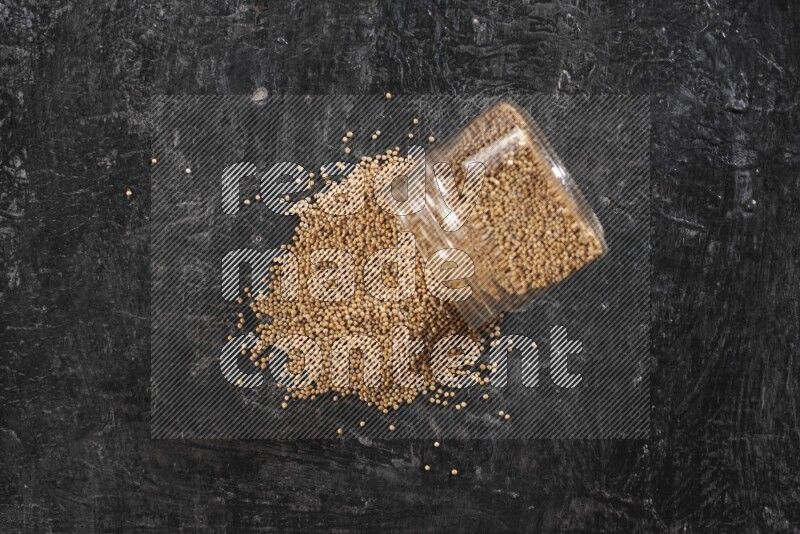 A glass jar full of mustard seeds and jar is flipped and seeds spread out on a textured black flooring