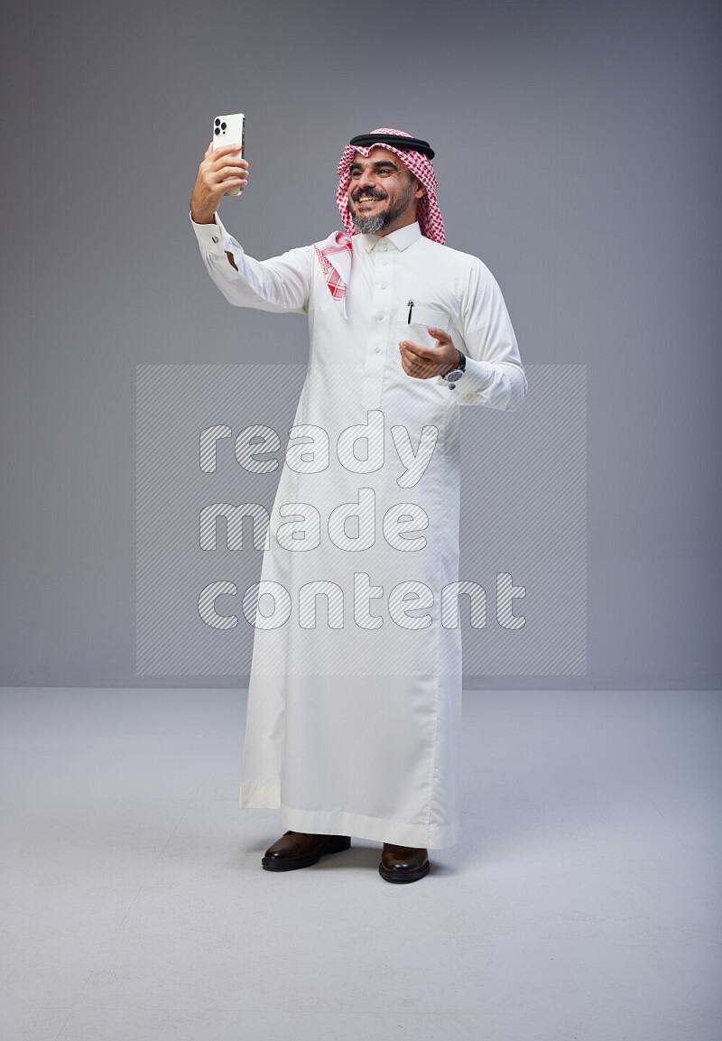 Saudi man Wearing Thob and red Shomag standing taking selfie on Gray background