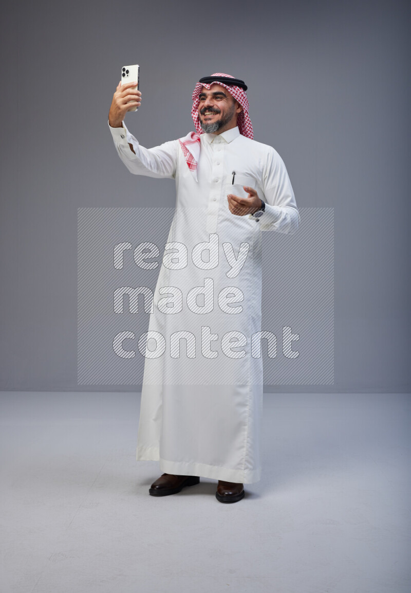 Saudi man Wearing Thob and red Shomag standing taking selfie on Gray background