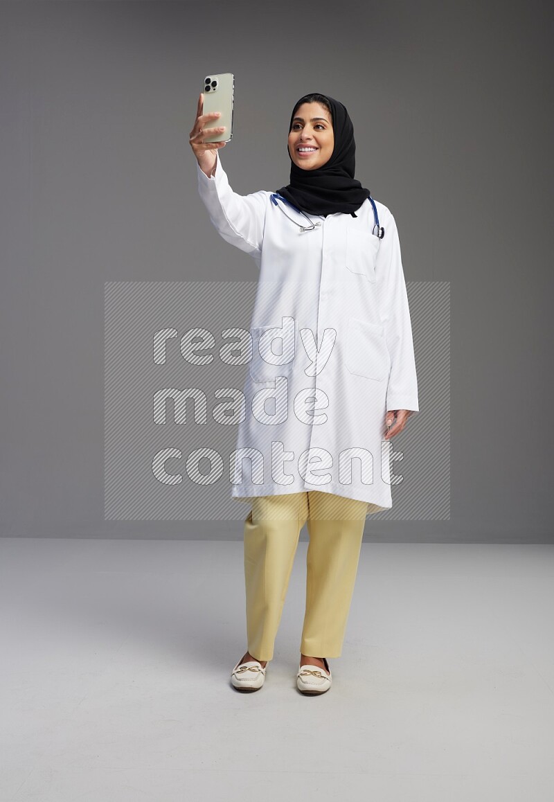 Saudi woman wearing lab coat with stethoscope standing taking selfie on Gray background