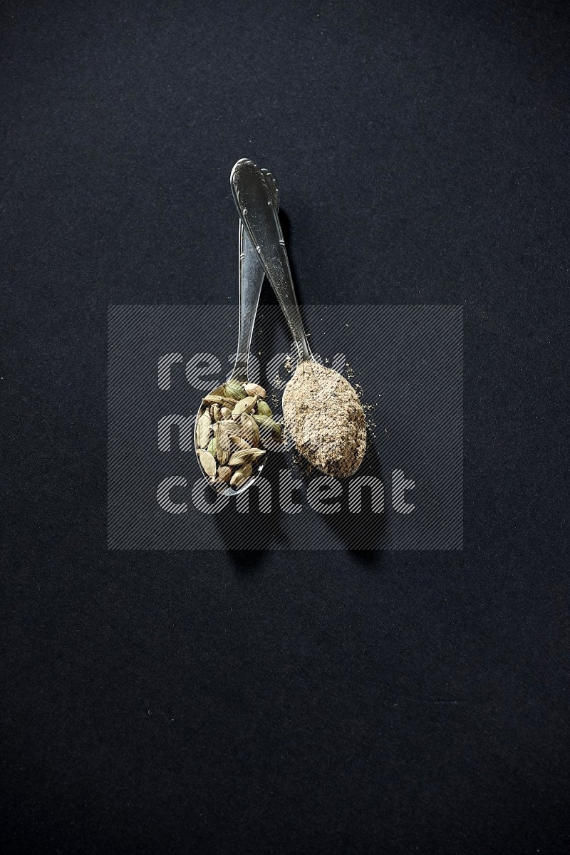2 Metal spoons full of cardamom powder and cardamom seeds on black flooring