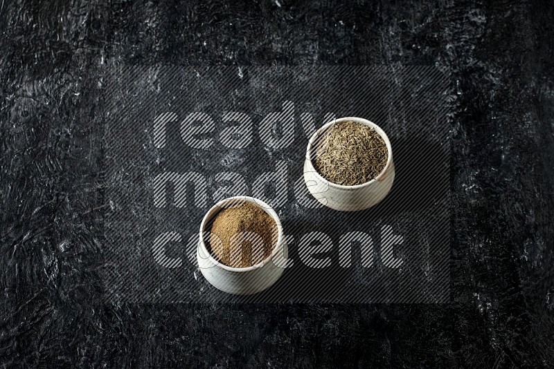 2 beige bowls, one full of cumin powder and the other full of seeds on a textured black flooring