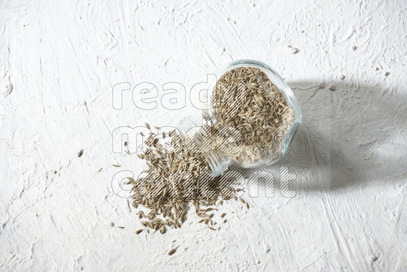 A flipped glass spice jar full of cumin seeds and the seeds spilled out on textured white flooring