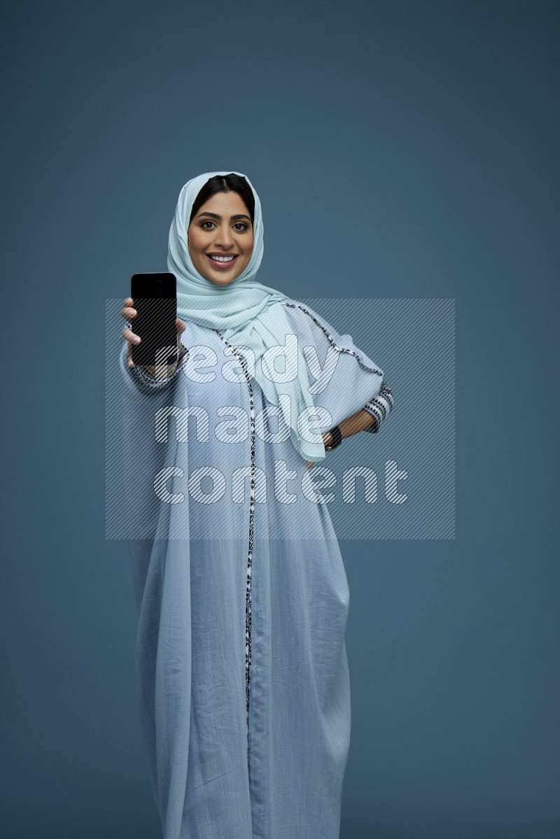 A Saudi woman Showing her phone screen in a blue background wearing a blue Abaya with hijab