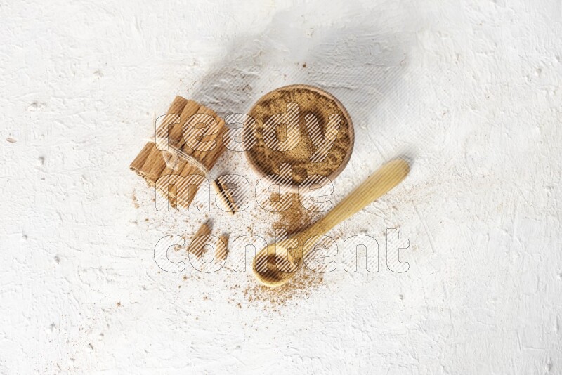 Cinnamon sticks stacked and bounded beside a wooden bowl full of cinnamon powder and a wooden spoon full of powder on white background