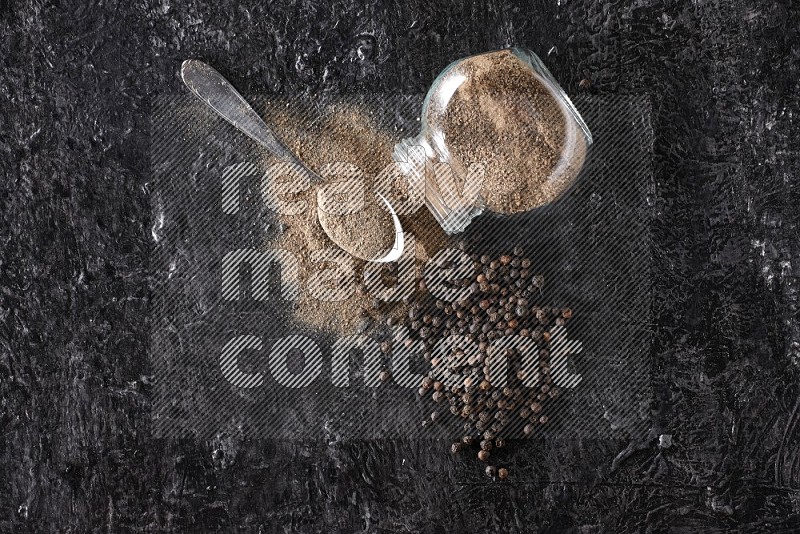 A flipped glass spice jar full of black pepper powder with a metal spoon full of powder and pepper beads on textured black flooring