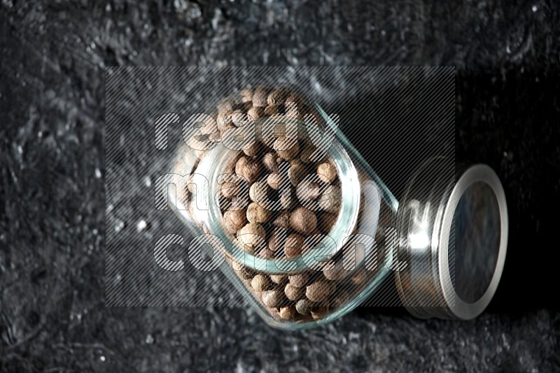 A glass spice jar full of allspice whole balls on a textured black flooring