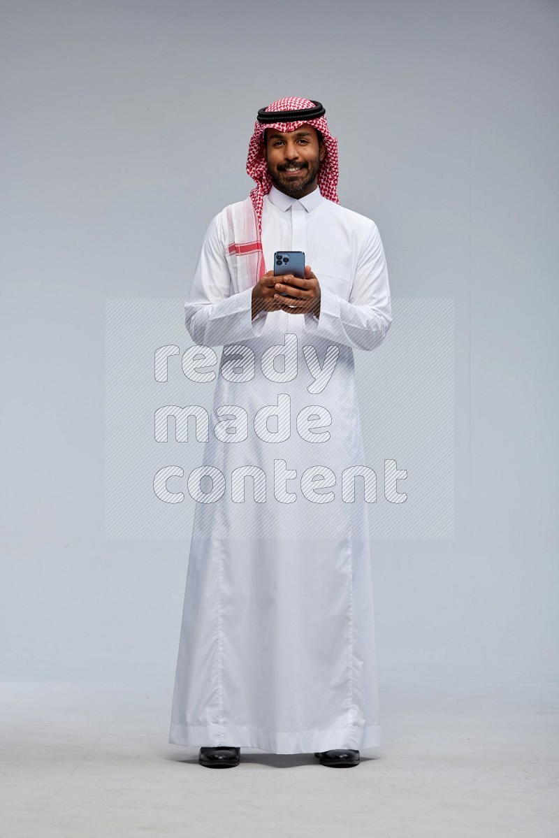 Saudi man Wearing Thob and shomag standing texting on phone on Gray background