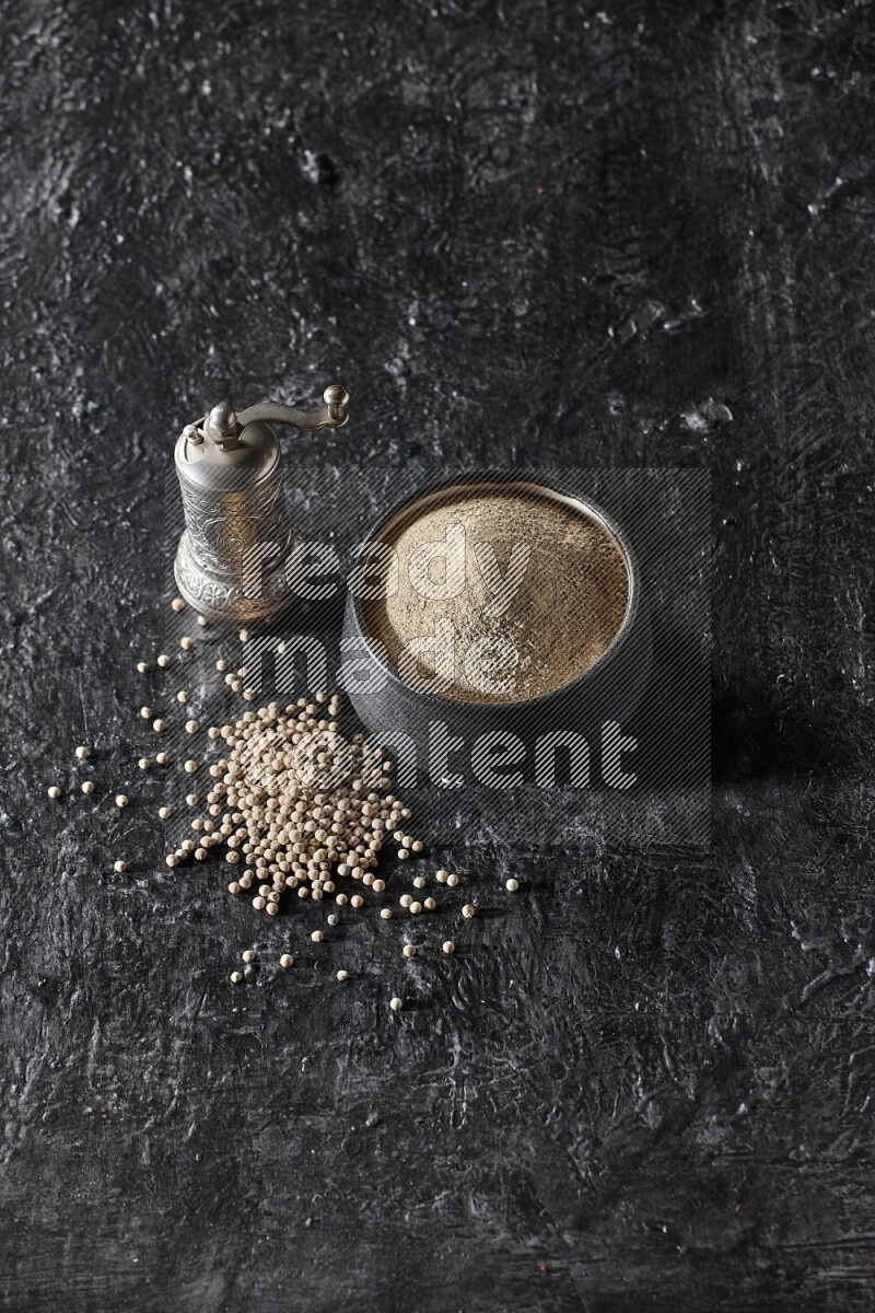 Black pottery bowl full of white pepper powder with pepper beads and metal pepper grinder on textured black flooring