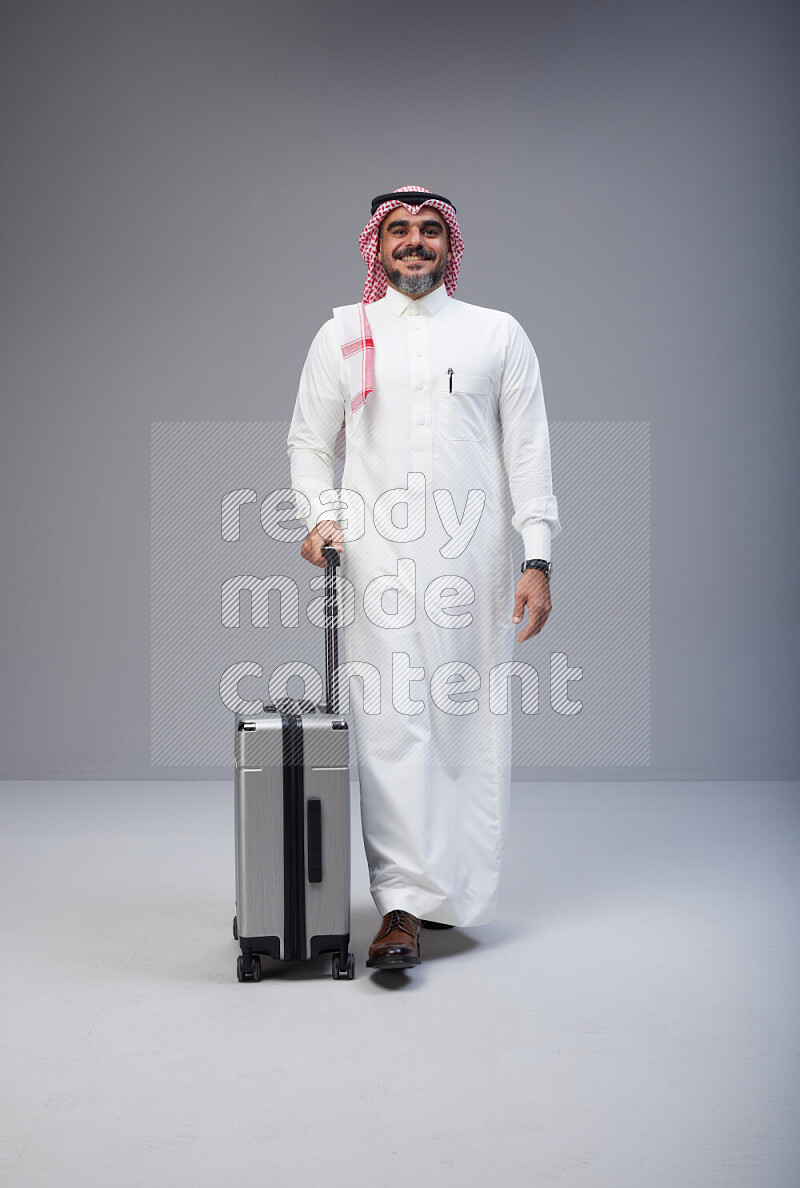 Saudi man wearing Thob and red Shomag standing holding Travel bag on Gray background