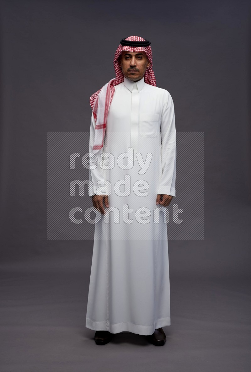 Saudi man wearing thob and shomag standing interacting with the camera on gray background