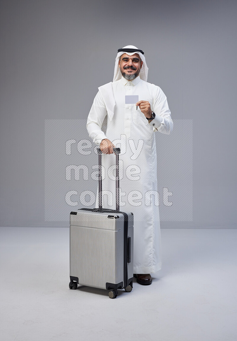 Saudi man wearing Thob and white Shomag standing holding Travel bag and ATM card on Gray background