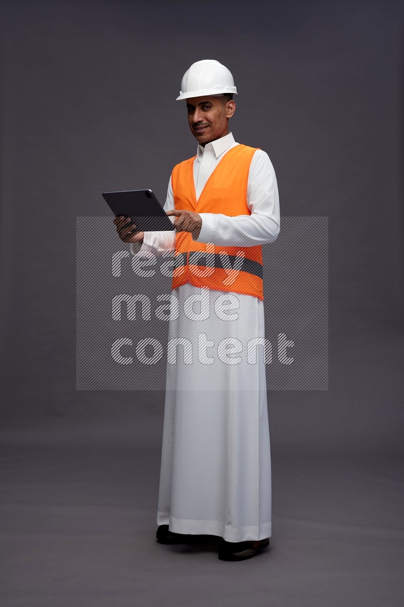 Saudi man wearing thob with engineer vest standing working on tablet on gray background
