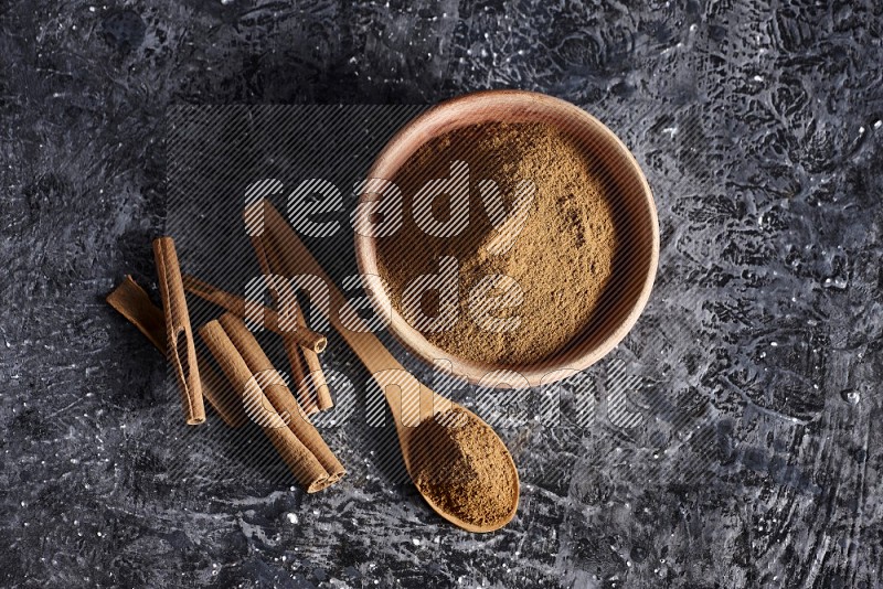 wooden bowl full of cinnamon powder and a wooden spoon full of it with cinnamon sticks on a textured black background