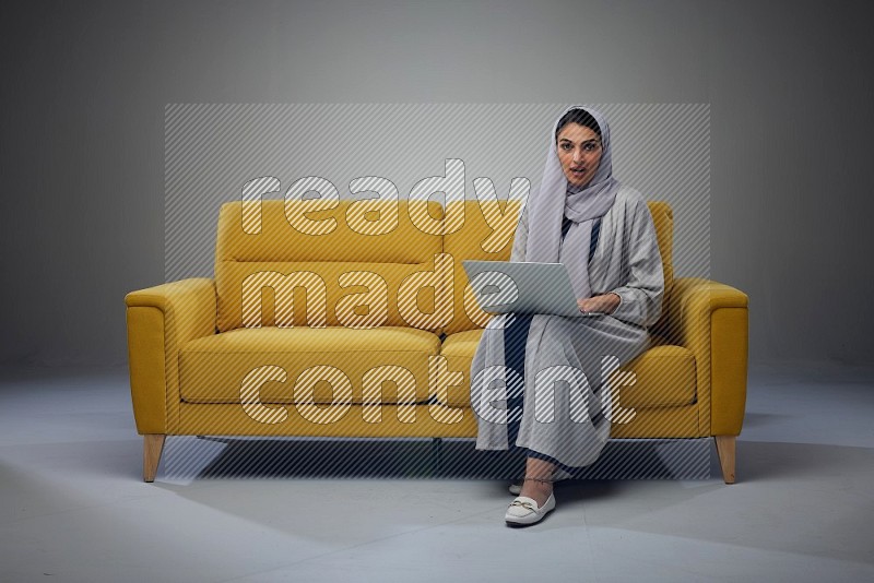 A Saudi woman wearing a light gray Abaya and head scarf sitting on a yellow sofa and using her laptop eye level on a grey background