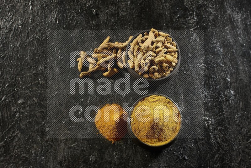 2 pottery black bowls, one full of turmeric powder and the other full of dried turmeric whole fingers on a textured black flooring