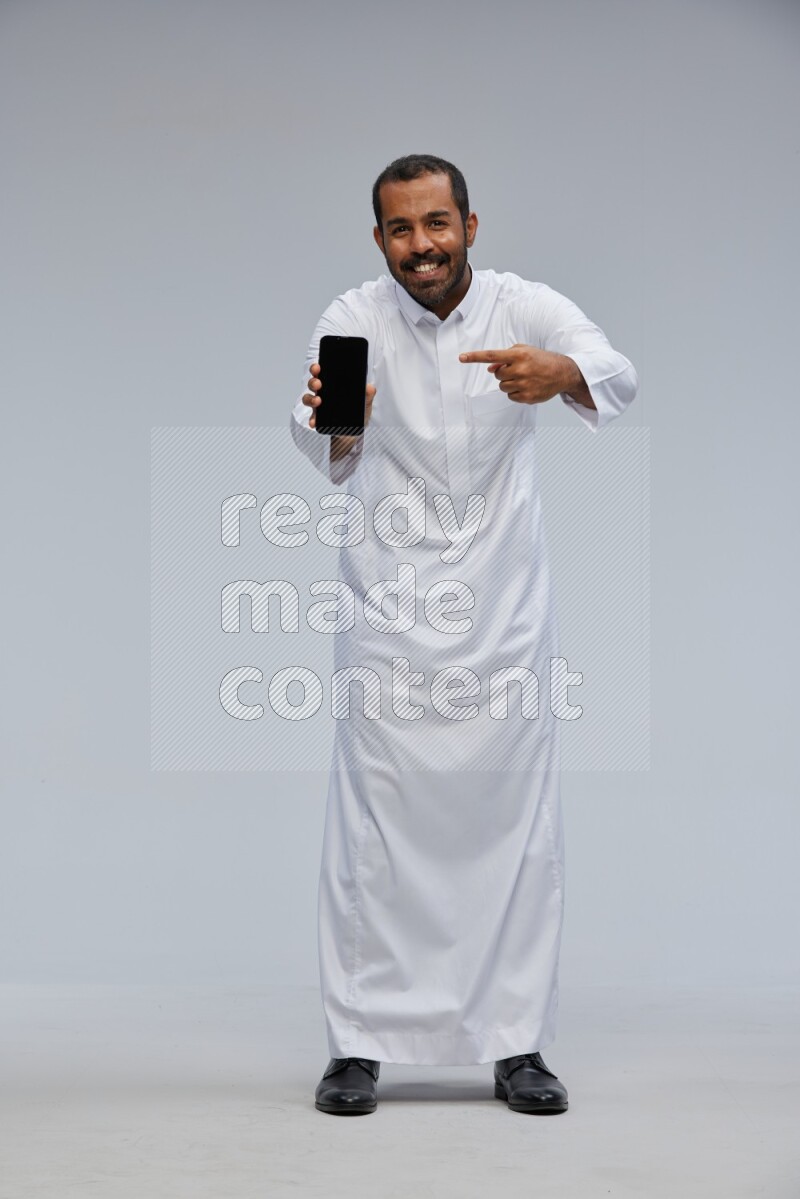 Saudi man wearing Thob standing showing phone to camera on Gray background