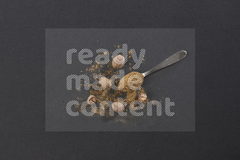 A metal spoon full of nutmeg powder with nutmeg powder and whole seeds beside it on a black flooring