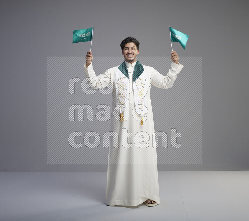 A Saudi man standing wearing thob and saudi flag scarf and holding small saudi flag on gray background