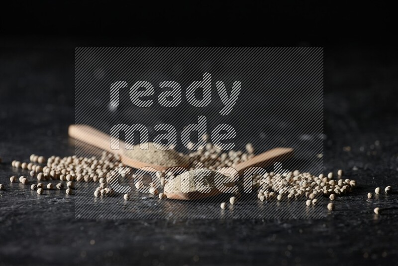 2 wooden spoons full of white pepper powder with white pepper beads on textured black flooring