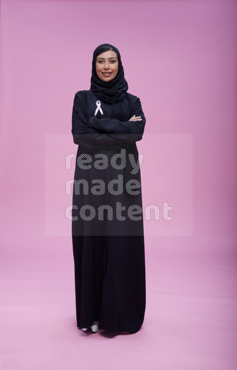 Saudi woman wearing pink ribbon on Abaya standing crossed arms on pink background