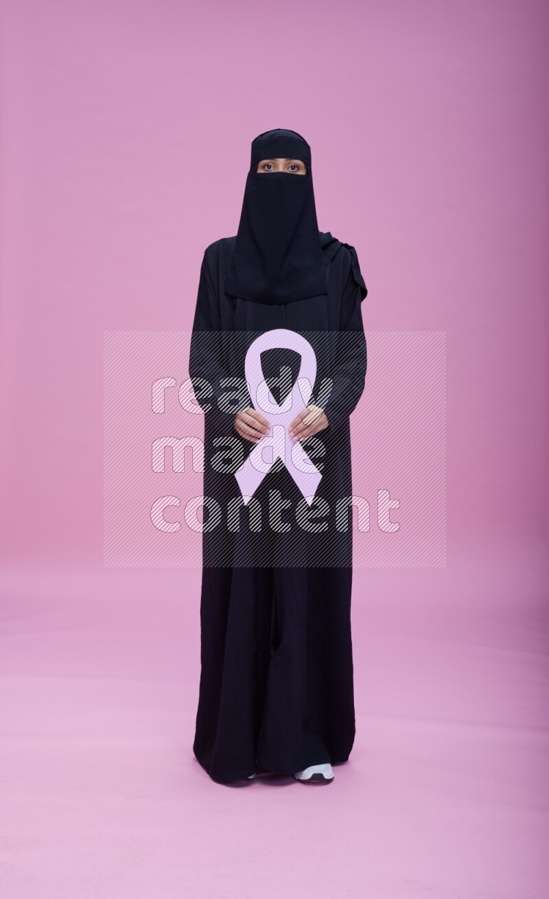 Saudi woman wearing abaya and niqab standing holding awareness ribbon on pink background