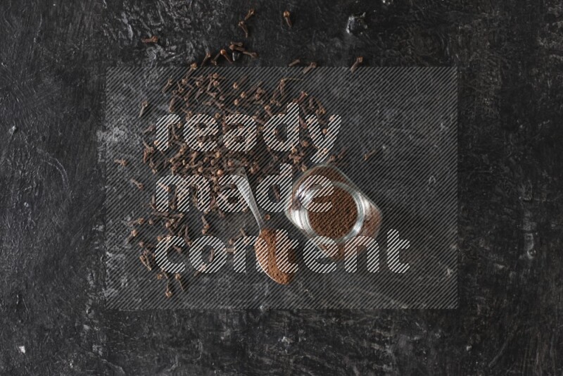 وعاء زجاجي للبهارات وملعقة معدنية ممتلئان ببودرة القرنفل مع حبوب القرنفل المتناثرة على أرضية سوداء