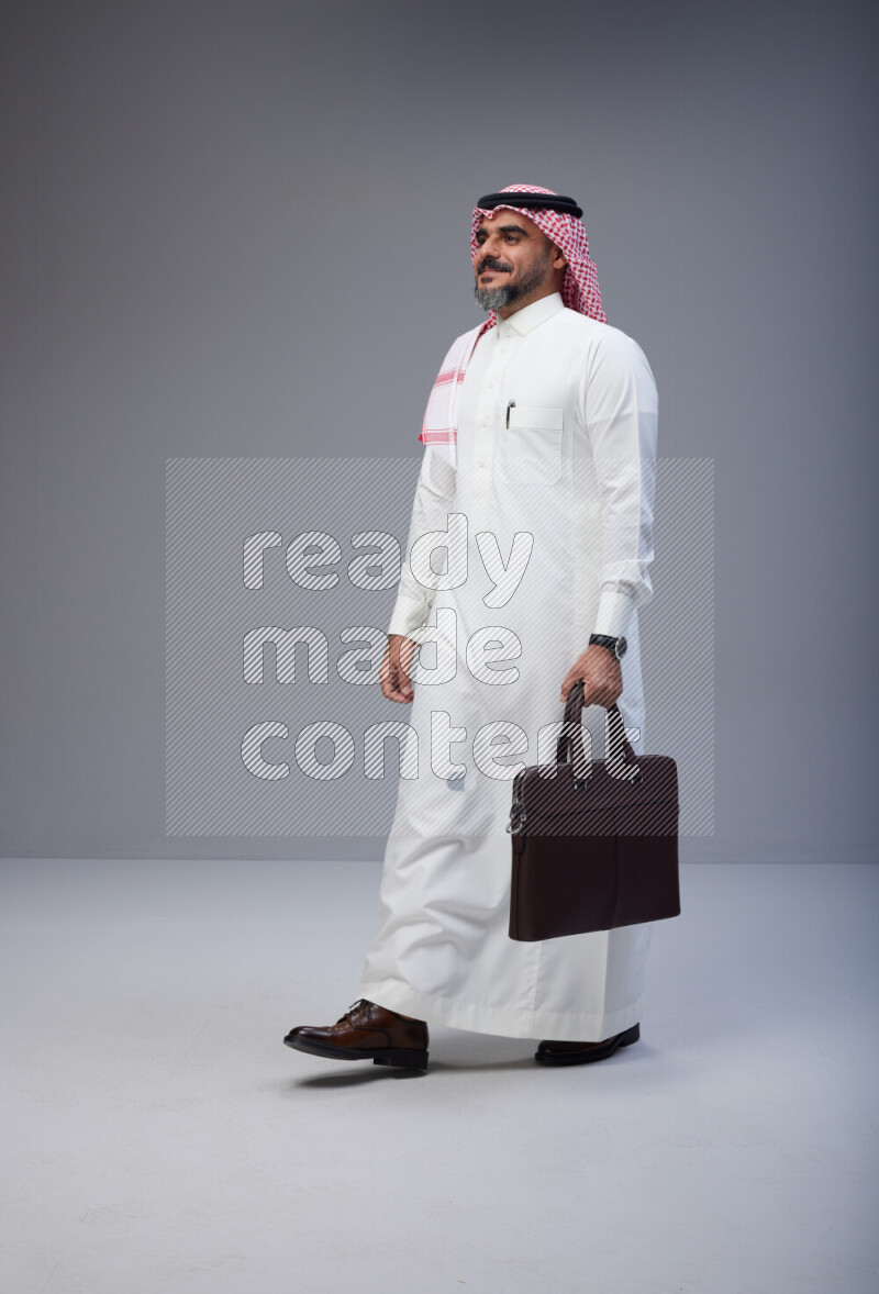 Saudi man Wearing Thob and red Shomag standing holding bag on Gray background