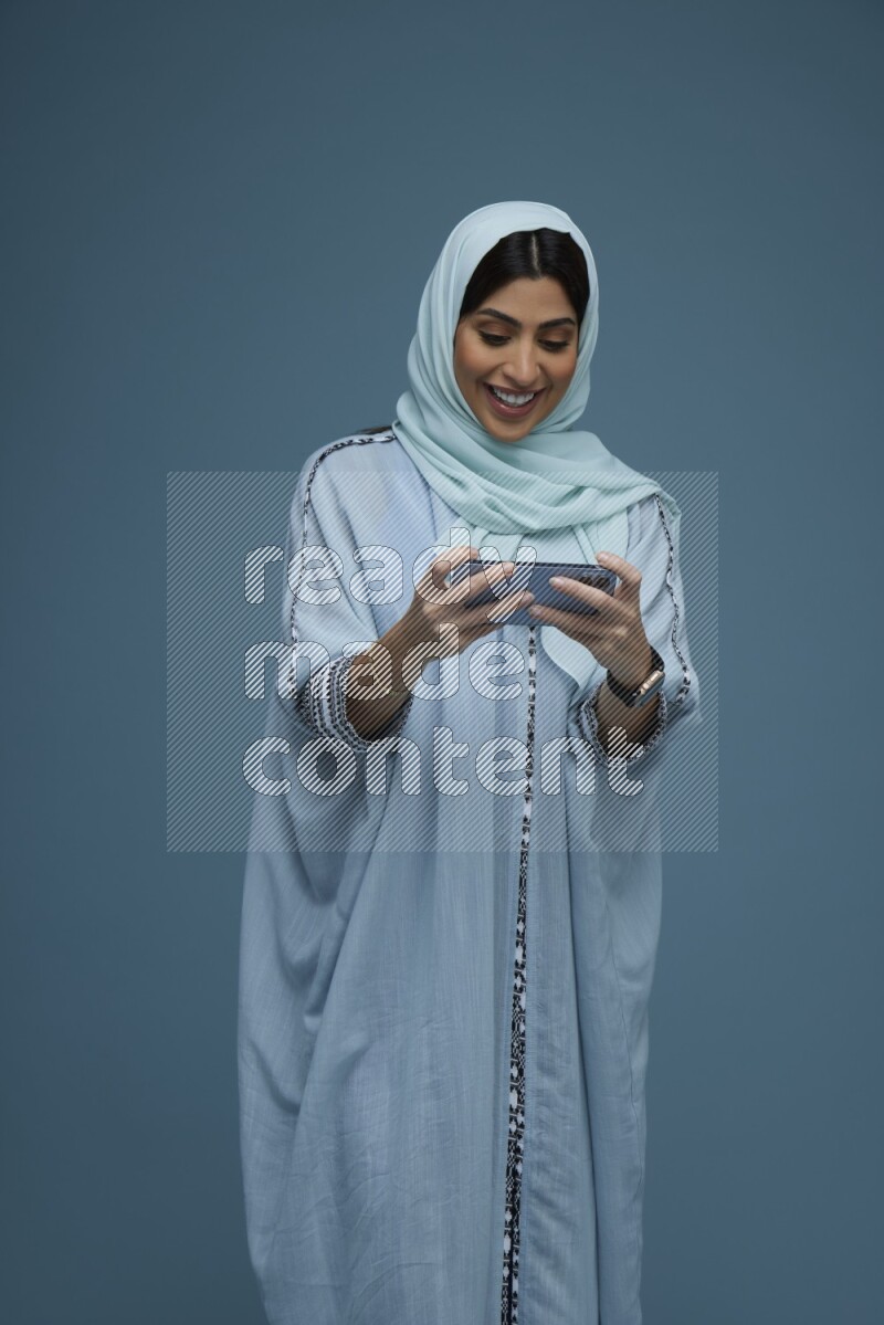 A Saudi woman Playing a Game on her phone on a blue background wearing a blue Abaya with hijab