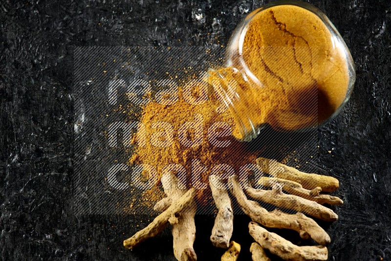 A flipped glass spice jar full of turmeric powder and powder spilled out of it with dried whole fingers on textured black flooring