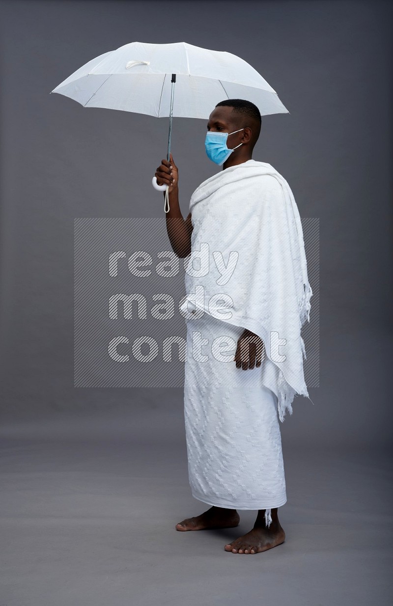 A man wearing Ehram with face mask Standing holding umbrella on gray background